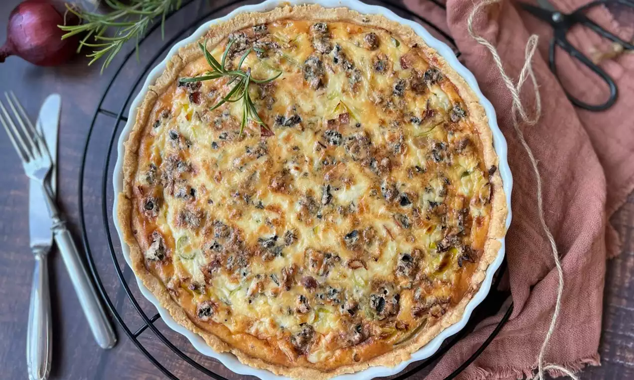 Slaný koláč zlatavý a křupavý, s nesouměrnou hnědou vrstvou slaniny a pórku, servírovaný v bílé keramické formě na drátěném podnosu