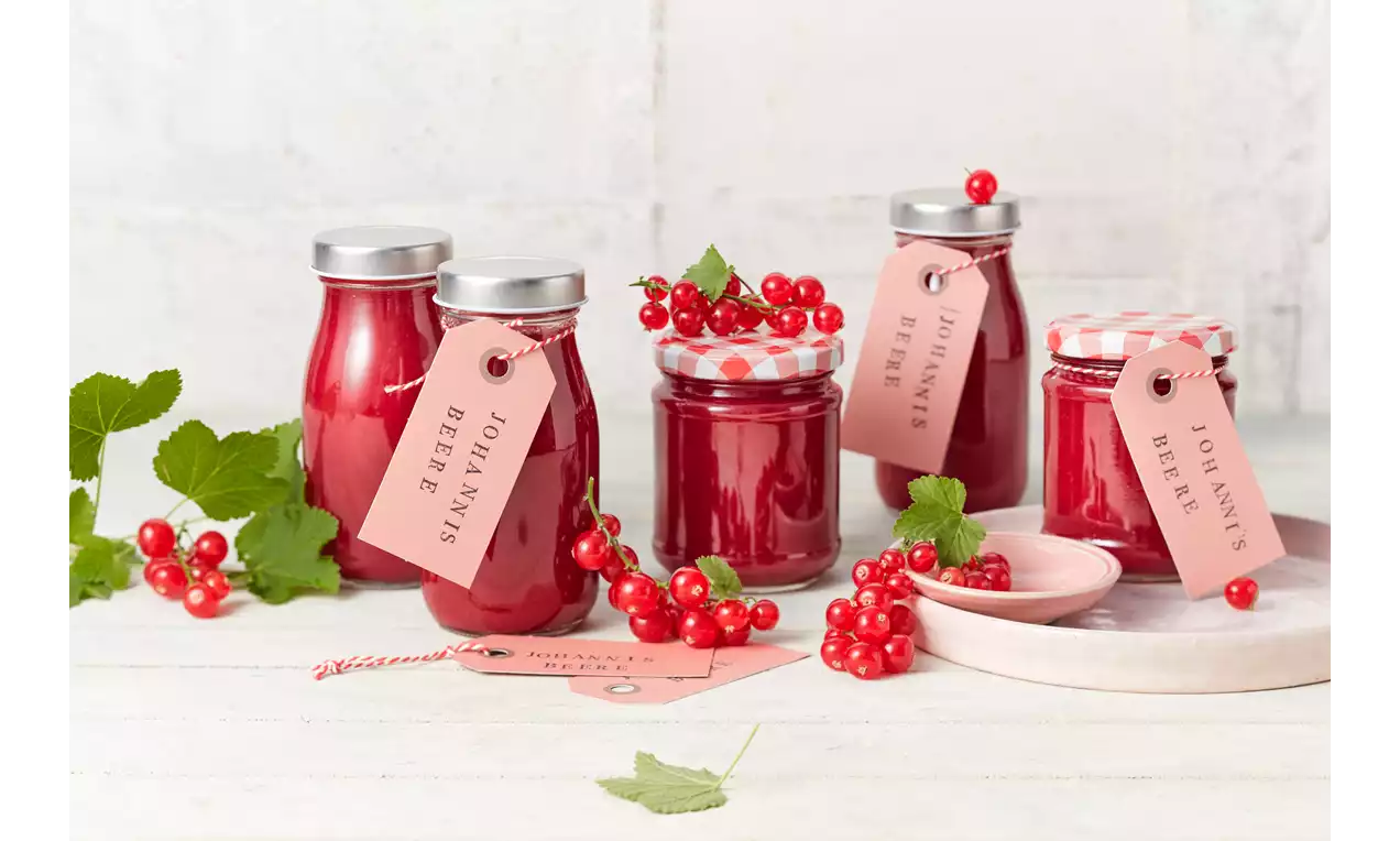 Glasbehälter mit tiefroter Marmelade, teils mit karierten Deckeln, daneben frische Johannisbeeren, Anhänger und ein rosafarbener Teller