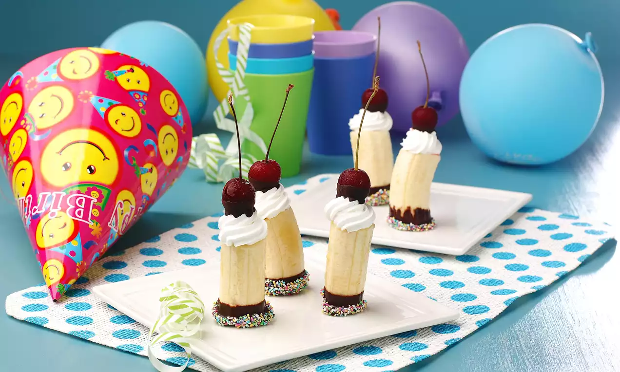 Renkli balonlar ve parti şapkasıyla çevrili beyaz tabakta, uçları çikolatalı ve renkli şekerli muzlar, krem şanti ve vişne ile süslenmiş