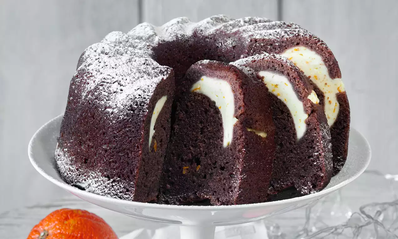 Kakaová bábovka tmavohnedá s krémovou tvarohovou náplňou a jemnými pomarančovými tónmi, posypaná práškovým cukrom na bielom podnose