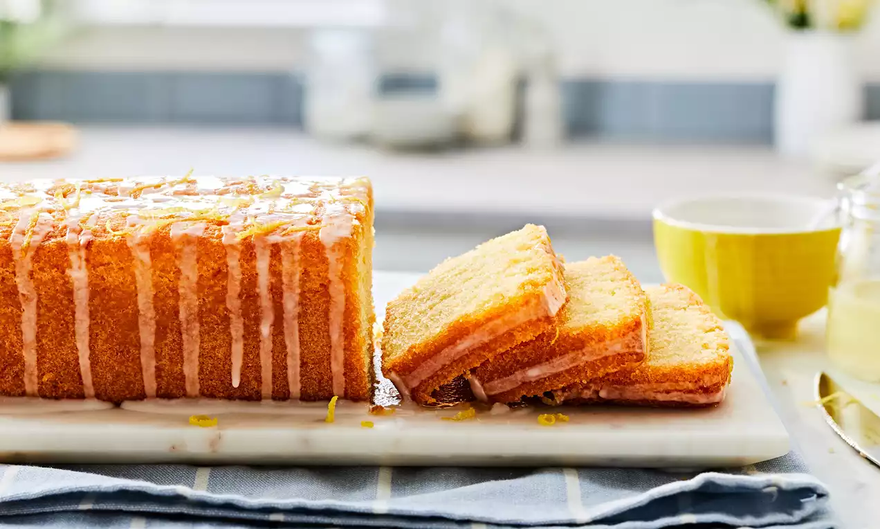Golden rectangular loaf cake with glossy lemon drizzle, textured slices arranged on a white marble board with lemon zest garnish