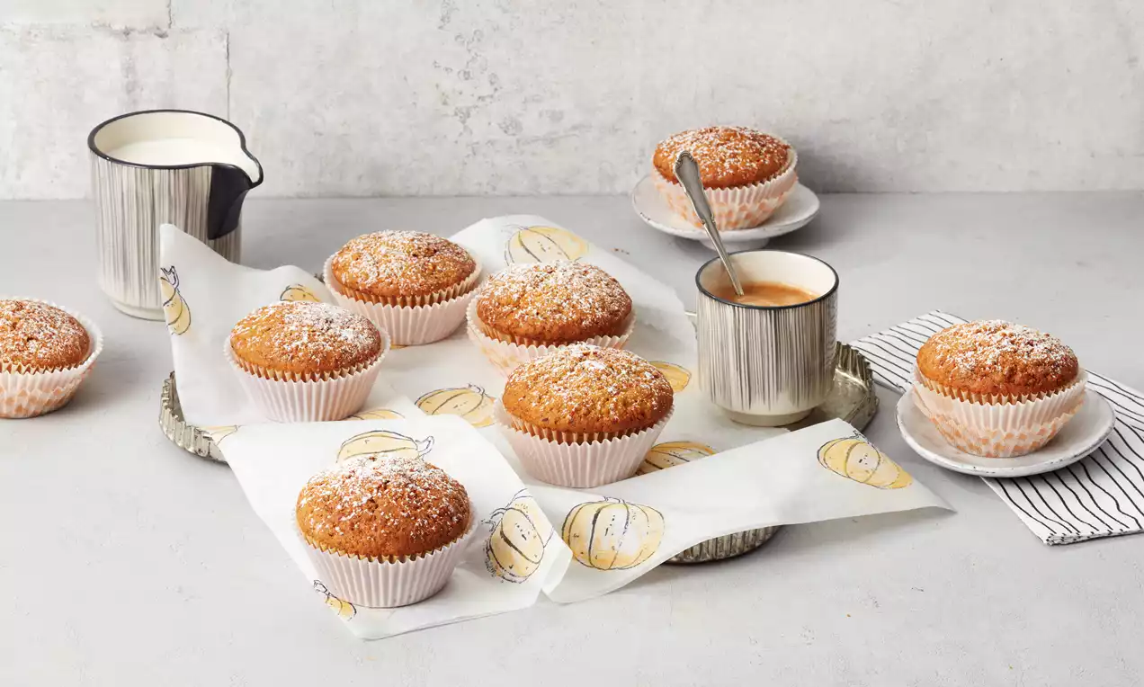 Goldbraune Kürbis-Muffins mit glatter Oberfläche und weißem Zuckerschnee in Papierförmchen auf herbstlich bedrucktem Backpapier angerichtet