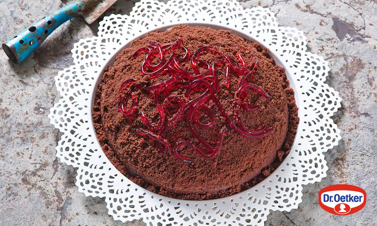 Okrúhly čokoládový tortový kopček posypaný tmavohnedými hoblinami, ozdobený červenými gumenými žížalami na papierovej čipke.