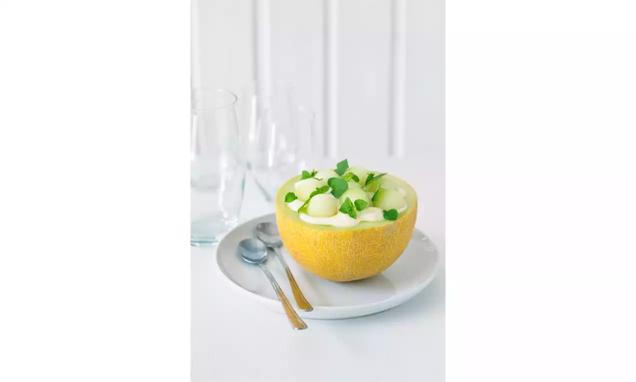 Polovica žltého melóna plnená krémovo bielym dezertom, ozdobená guľôčkami melóna a mätovými lístkami na bielom tanieri s lyžičkami