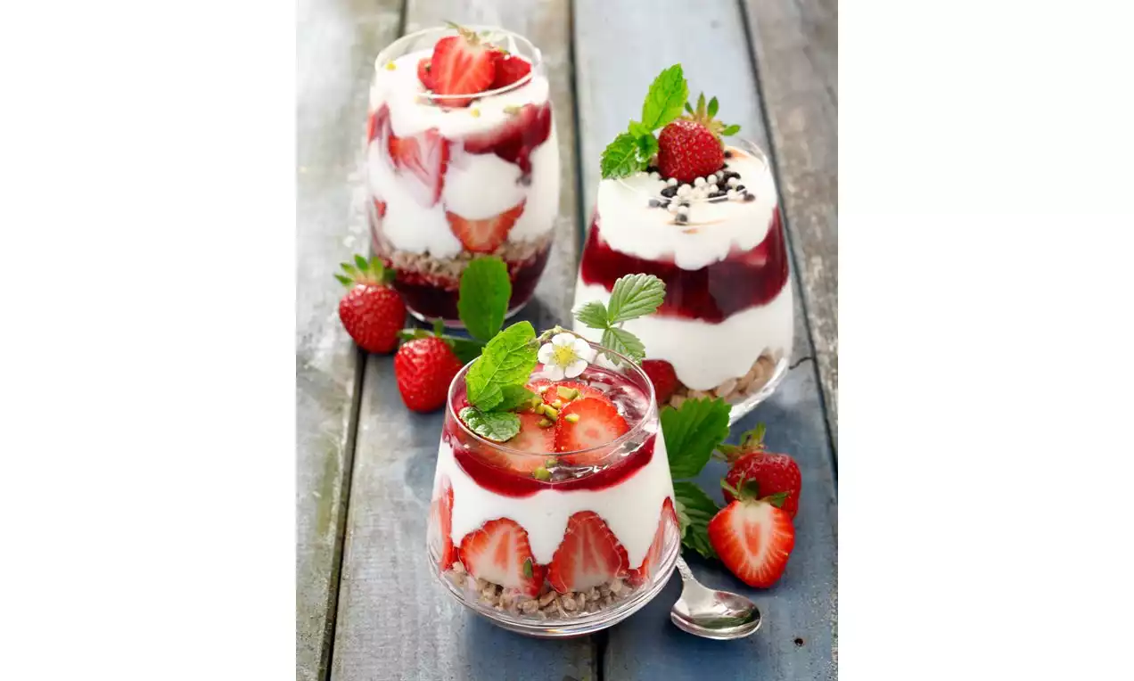 Drie glazen met laagjes romige yoghurt, rode aardbeien, rode coulis en granola, gegarneerd met muntbladeren en verse aardbeien