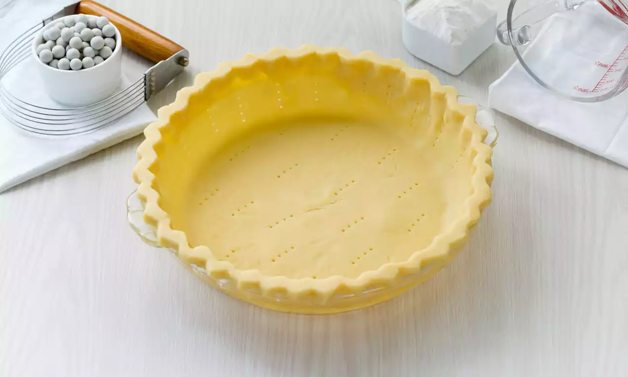Golden unbaked pie dough with scalloped edges pressed into a glass pie dish, pierced with fork marks, on a white surface with baking tools