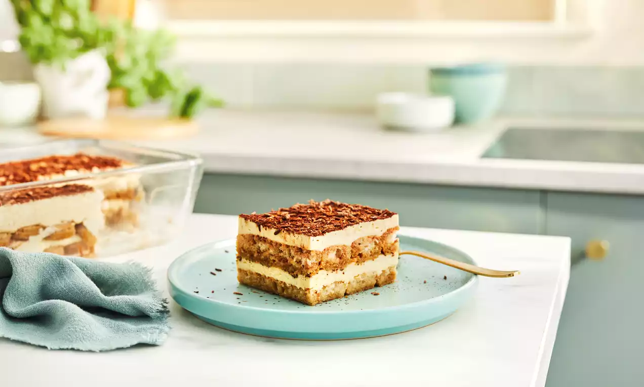 Layered tiramisu with pale cream and coffee-soaked sponge topped with fine cocoa dust on a light blue plate beside a gold spoon