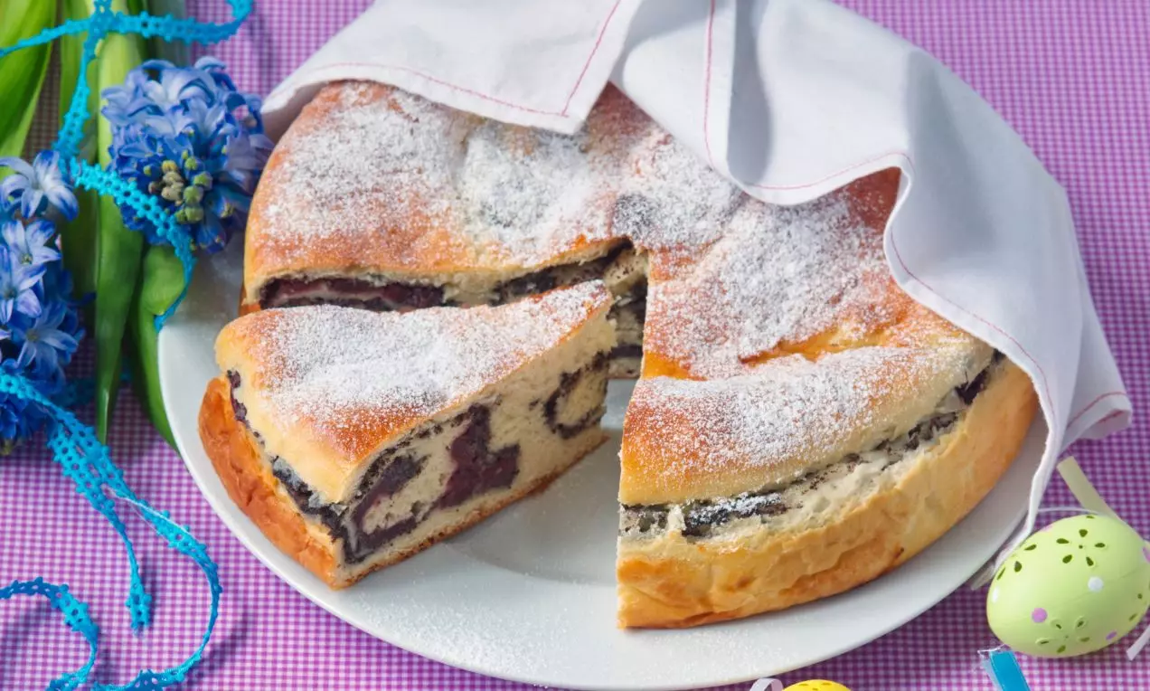 Mazanec světle zlatavé barvy s tmavými makovými pásy a višňovými kousky, lehce posypaný moučkovým cukrem na bílém talíři s dekorem květin