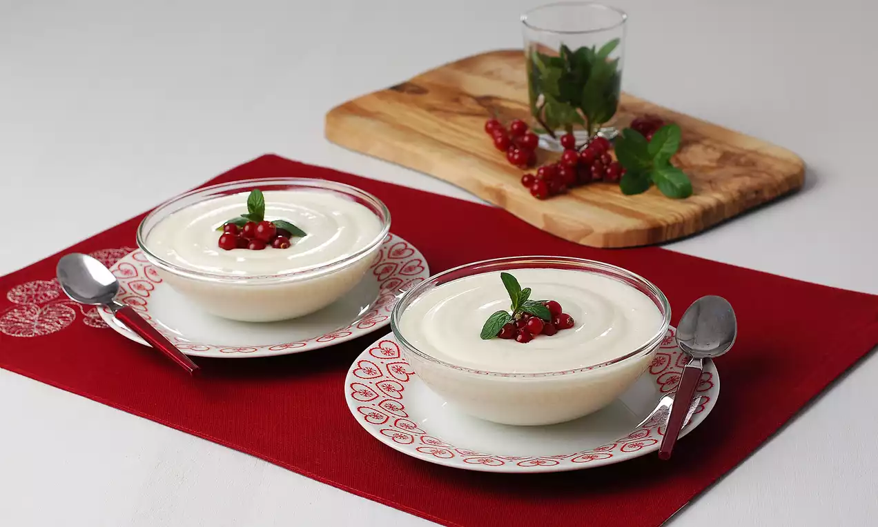 Kase içinde pürüzsüz beyaz muhallebi, üzeri kırmızı frenk üzümü ve yeşil nane yaprağı ile süslenmiş, kırmızı örtü üzerinde sunulmuş