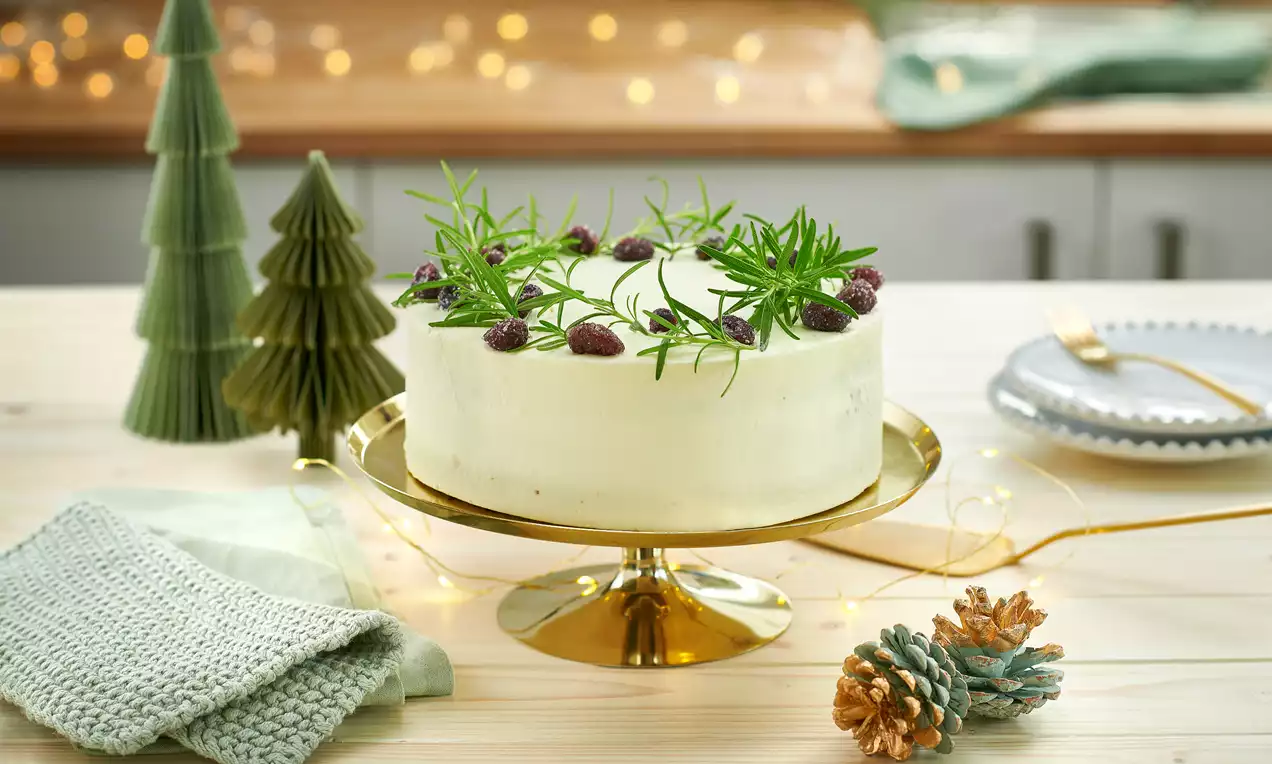 Weißer Adventskuchen mit glatter Oberfläche, dekoriert mit grünen Rosmarinzweigen und roten Beeren auf goldener Tortenplatte drapiert
