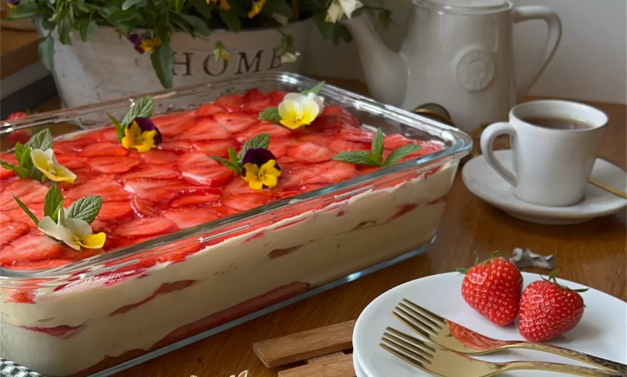 Jemné vrstvy krémového tiramisu v sklenenom pekáči, ozdobené jasne červenými plátkami jahôd, bylinkami a žltými kvetmi, na drevenom stole