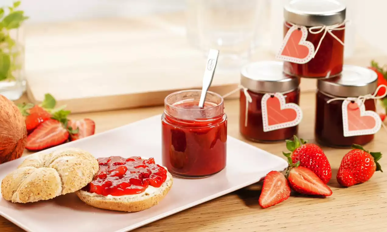 Sklenice jahodovo-kokosového džemu s lesklou červenou texturou na světlém stole vedle čerstvých jahod, pečiva a džemu na talíři