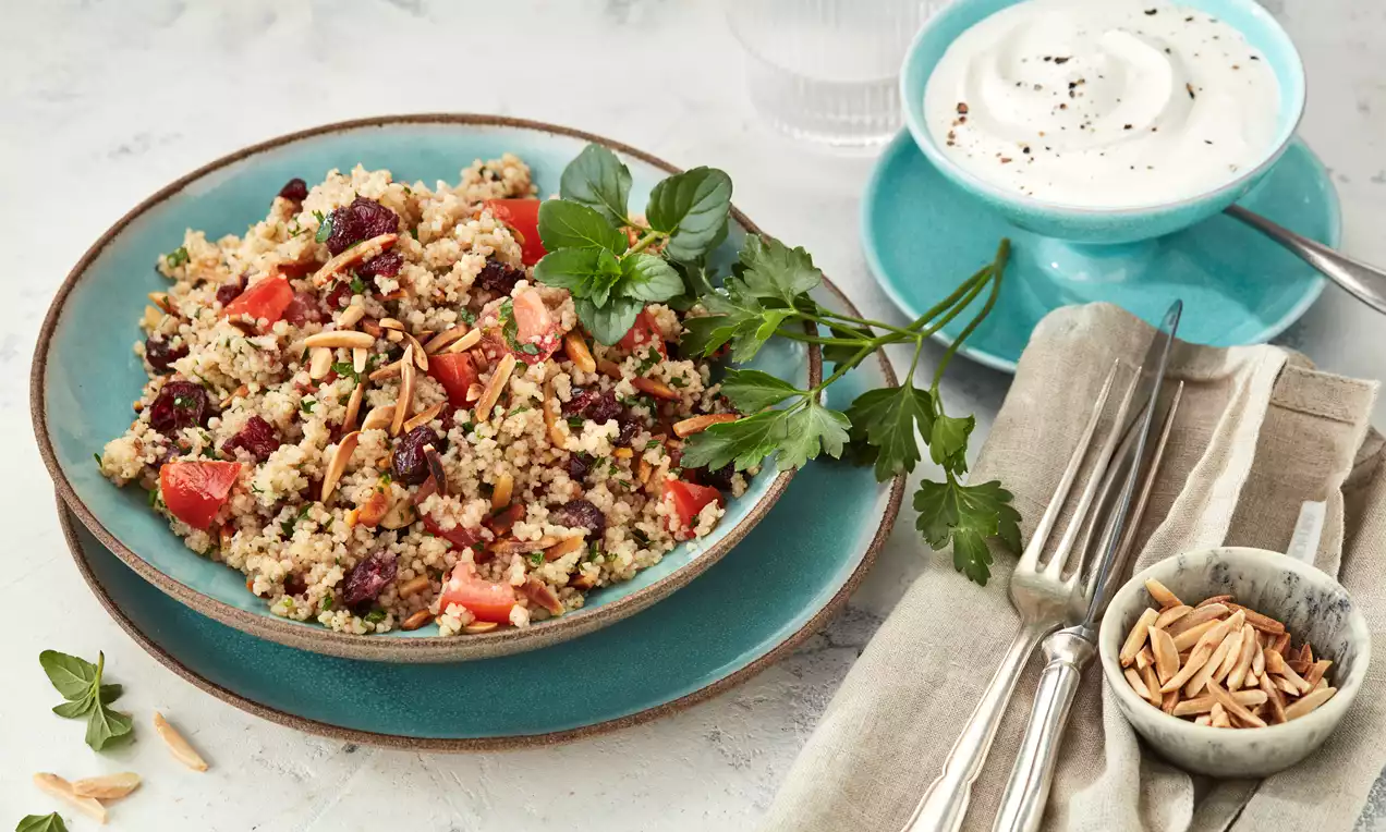 Bunter Couscous-Salat mit roten Tomatenstücken, frischen Kräutern und Mandelsplittern auf türkisfarbenem Teller, daneben Joghurt-Dip und Besteck