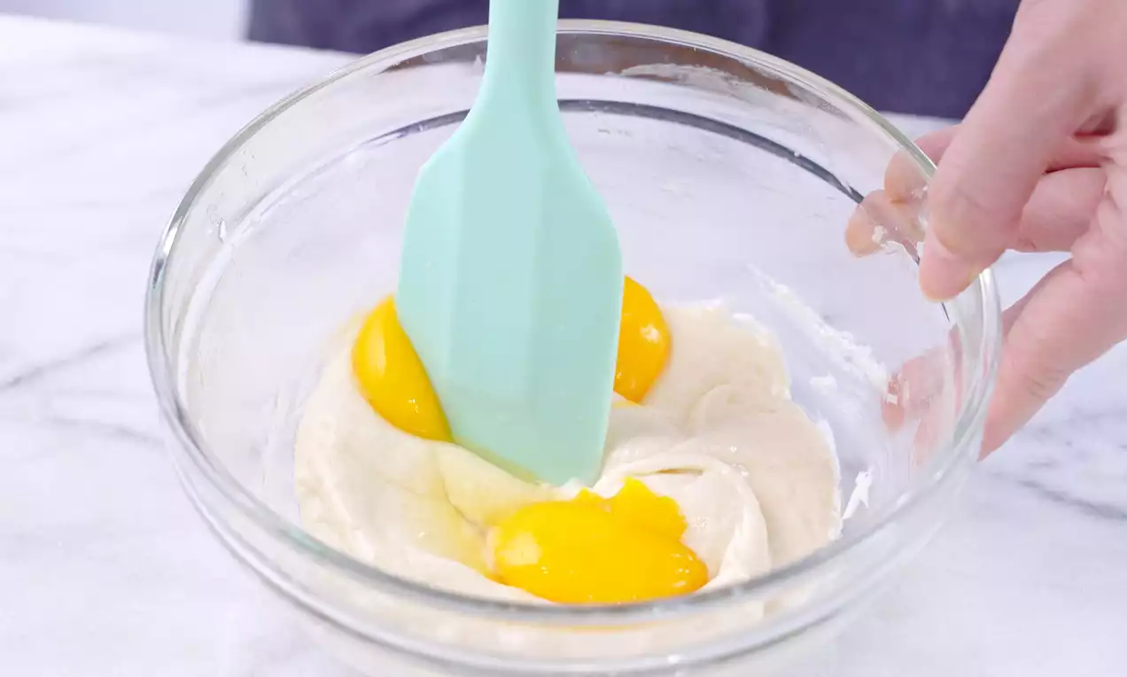 A mint-green silicone spatula stirring two bright yellow egg yolks into pale, creamy batter in a clear glass bowl on a marble surface