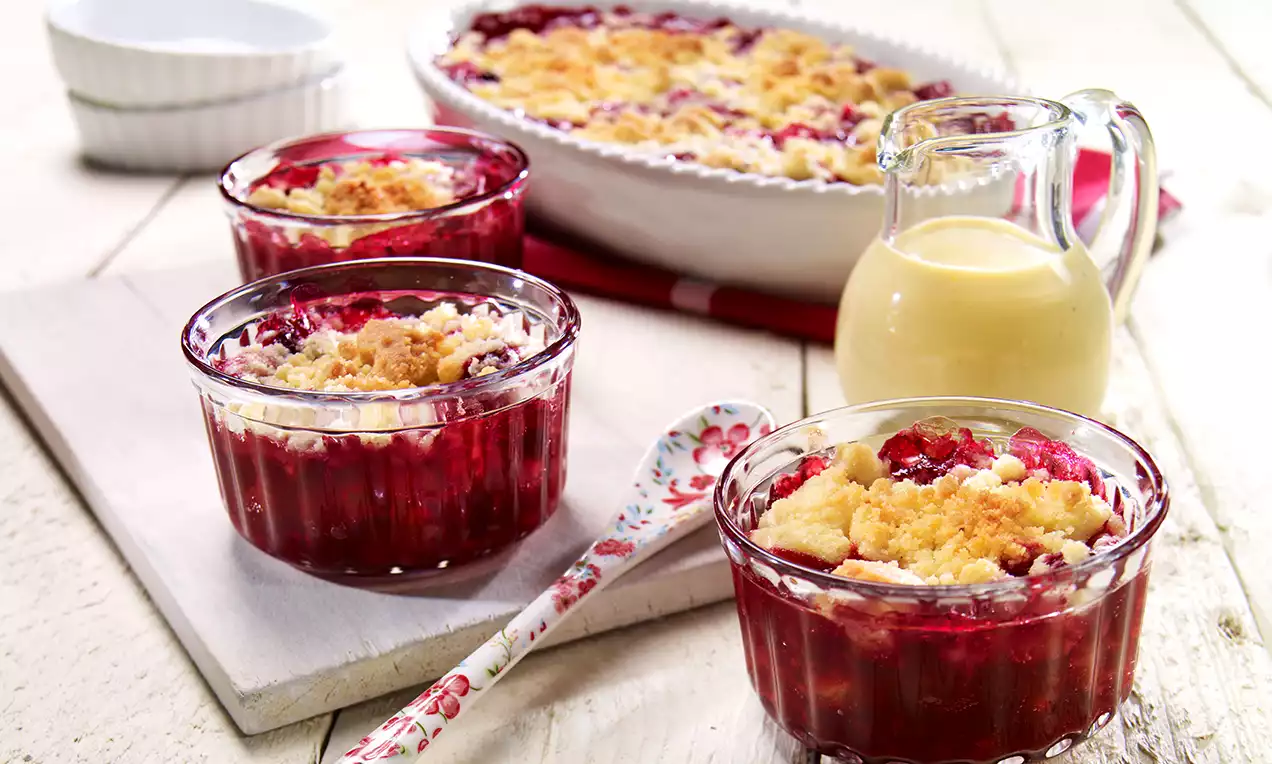 Rote Grütze in transparenten Dessertschalen mit goldbrauner Streuseldecke, daneben helle Vanillesoße in Glaskaraffe auf Holzbrett drapiert