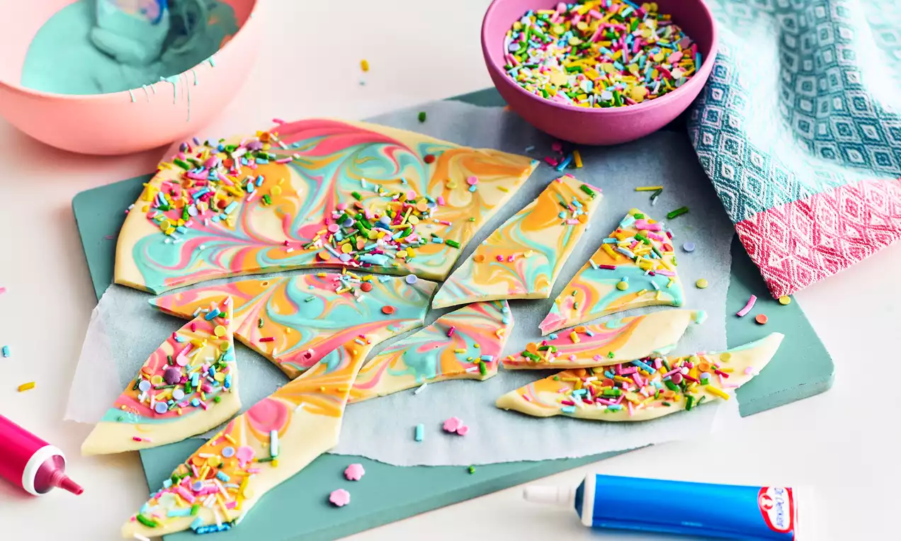 Colorful swirled chocolate shards with yellow, pink, blue, and green marbling, topped with bright rainbow sprinkles on parchment paper