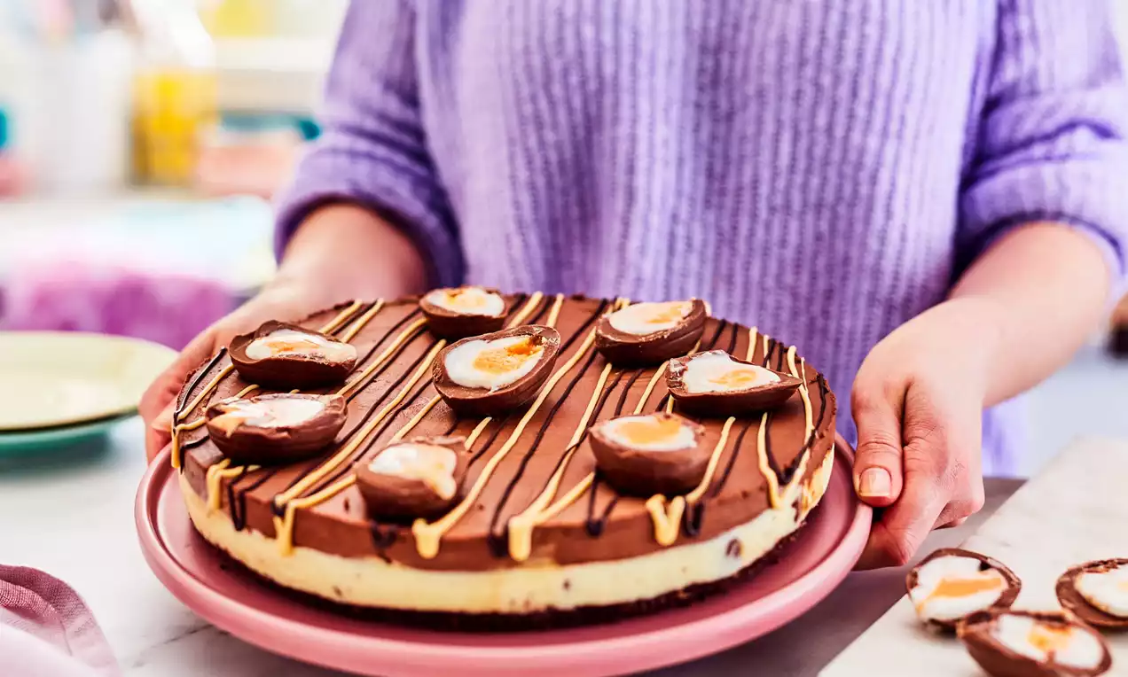 Creamy cheesecake with a pale yellow base and rich chocolate top, decorated with halved creme eggs and drizzled caramel lines on a pink plate