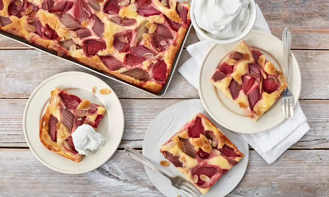Erdbeer-Rhabarber-Kuchen mit goldbrauner Kruste, roten Fruchtstücken und Mandelblättern auf Tellern mit Sahne, angerichtet auf Holzoptik-Untergrund
