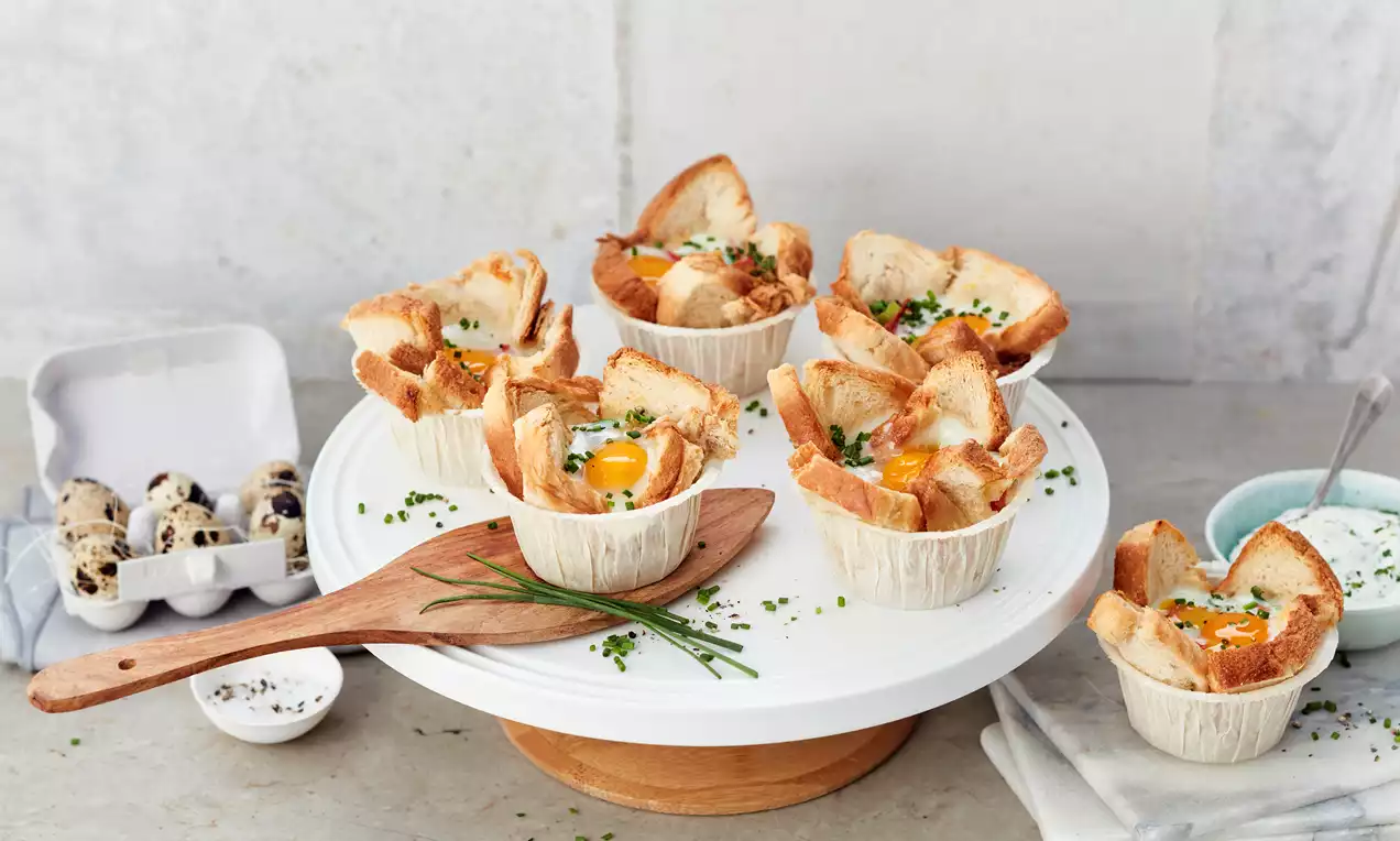 Muffins sărate aurii cu ou și pâine crocantă, servite în forme de hârtie, decorate cu cebuleț și așezate pe un platou alb