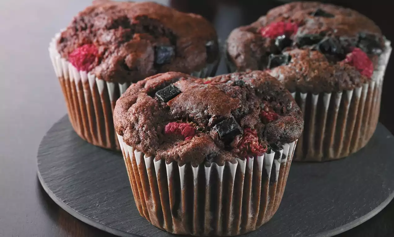 Three rich chocolate muffins with juicy raspberry pieces and dark chocolate chunks, set in white paper cups on a round slate plate