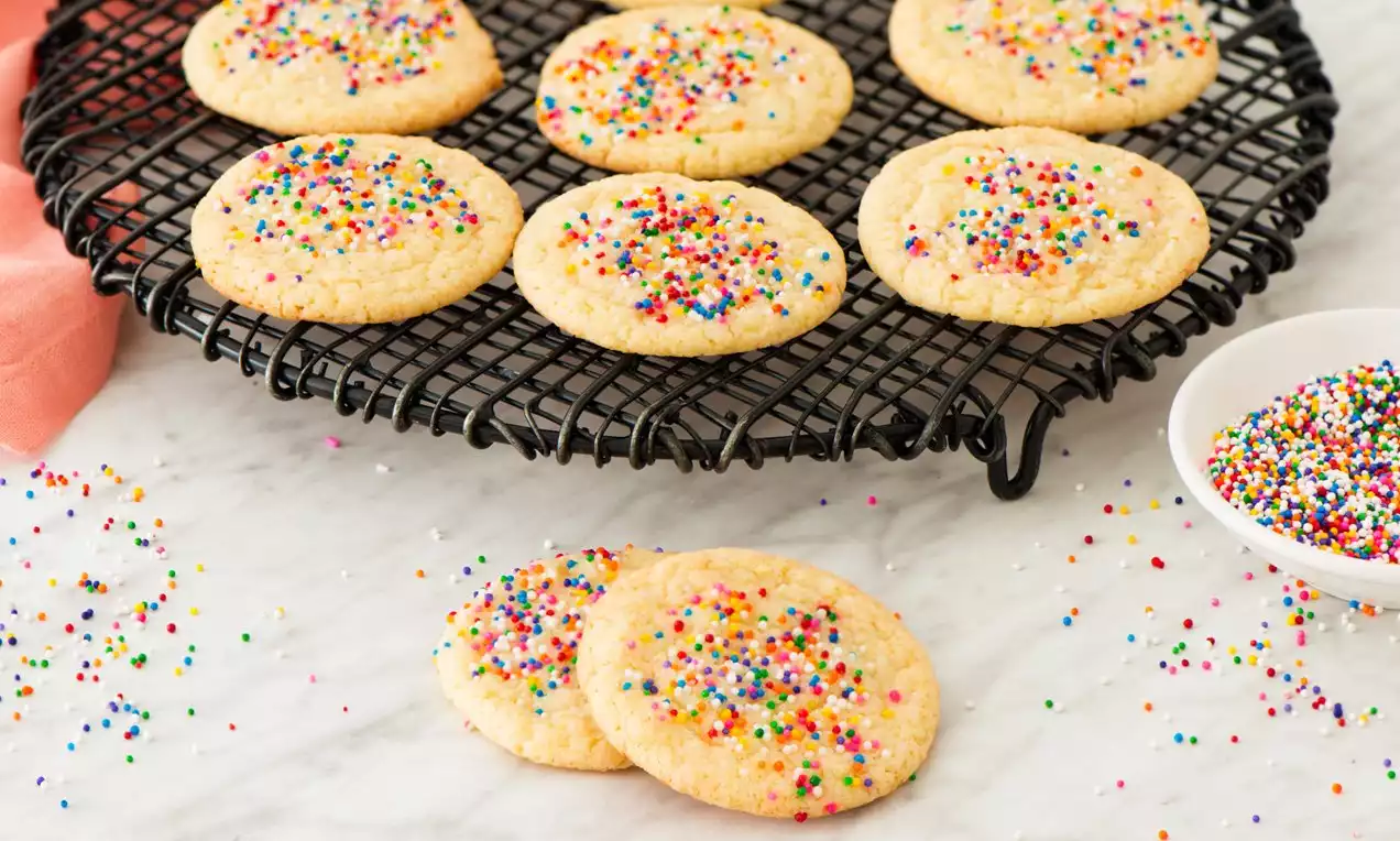Soft yellow cookies speckled with rainbow sprinkles on a black wire cooling rack, scattered sprinkles on marble with a white bowl nearby