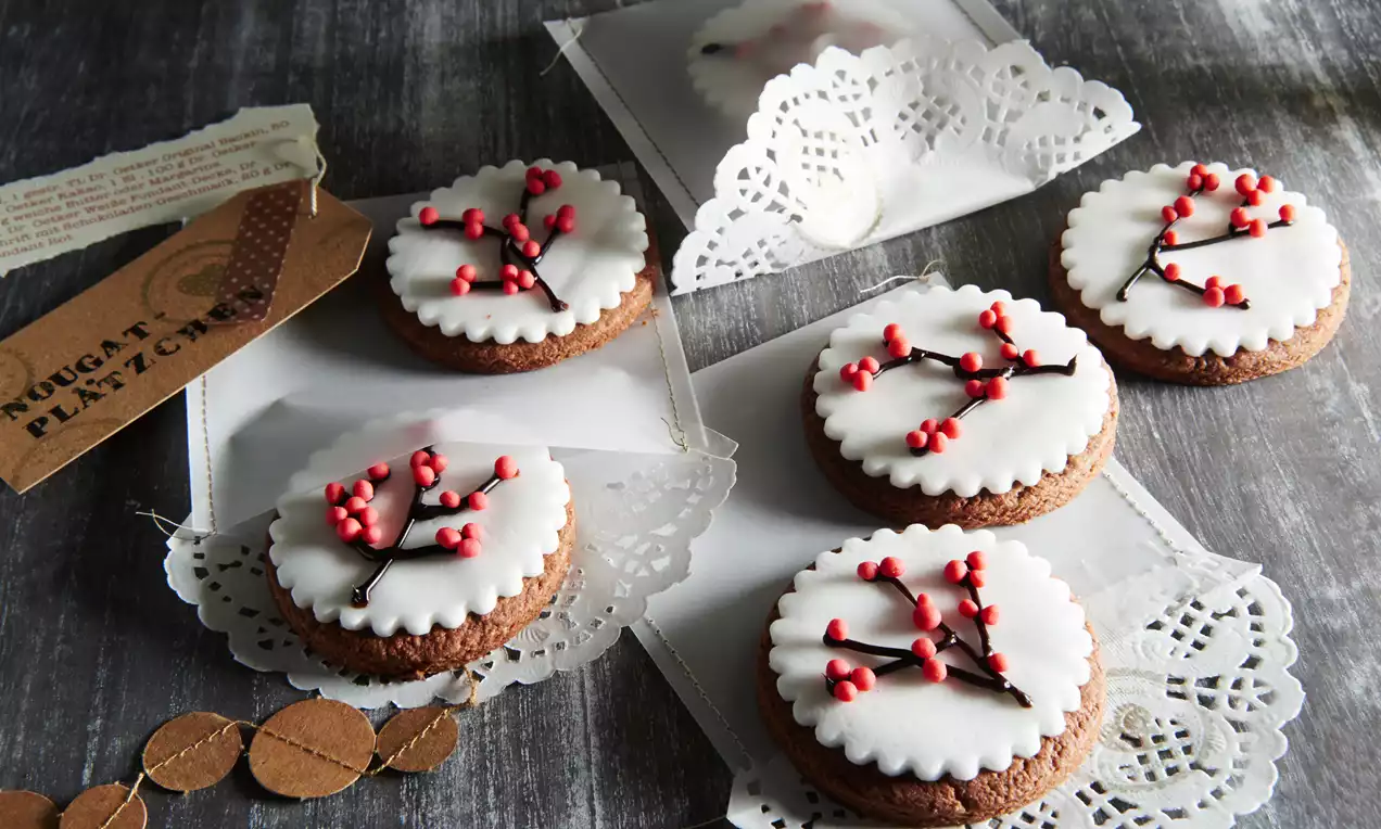 Runde Plätzchen mit glatter weißer Fondantdecke, verziert mit roten Beeren und schwarzen Ästen auf Spitze und grauem Untergrund arrangiert