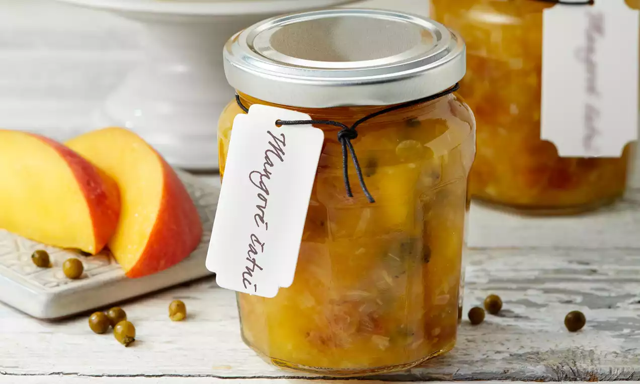 Mangové čatní v průhledné sklenici s kovovým víčkem, viditelné kousky ovoce a koření, vedle plátky manga na světlém dřevěném podkladu