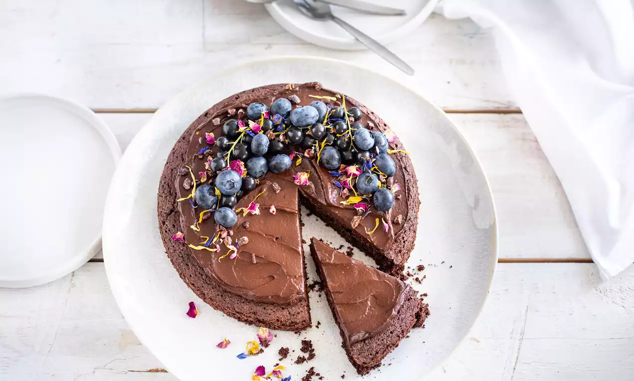 Dunkler, cremiger Schokoladenkuchen mit glänzender Oberfläche, dekoriert mit frischen Blaubeeren und essbaren Blüten auf weißem Teller