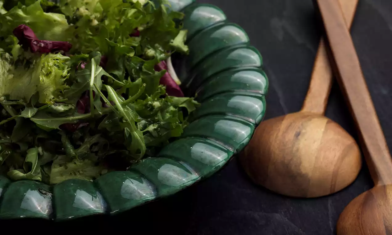 Grönsallad med skrynkliga salladsblad i olika gröna nyanser och inslag av mörkröda strimlor, upplagd i en djup grön skål på svart yta