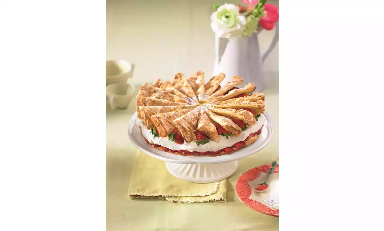 Koláč z listového těsta ve tvaru hvězdicového kruhu na bílém stojanu, vrstvy šlehačky a jahod, ozdobený květinami v pozadí