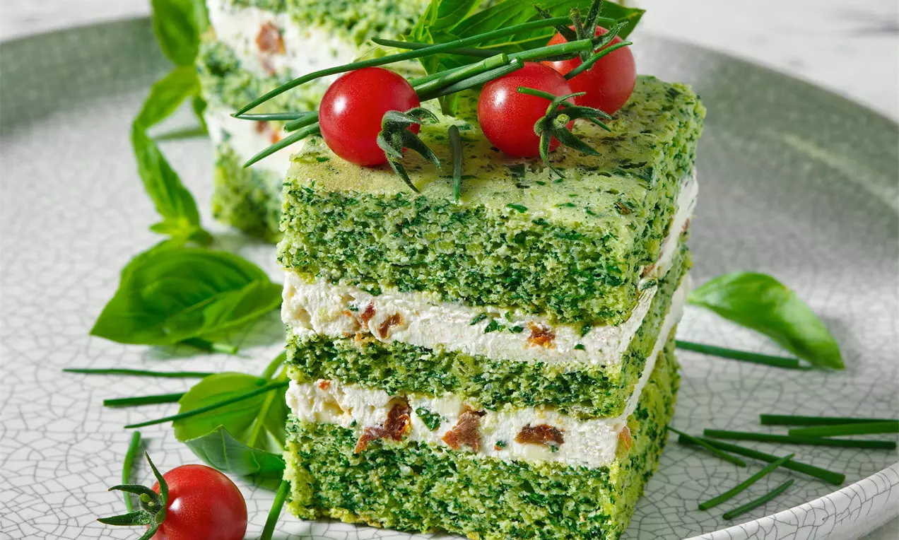 Trojvrstvé špenátové řezy s bílou sýrovou náplní, zdobené čerstvými cherry rajčaty, pažitkou a bazalkou na světle šedém talíři