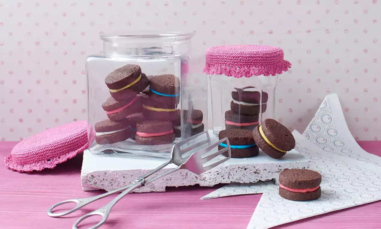 Čokoládové sušienky s jasne farebnou krémovou náplňou v pohároch s ružovým háčkovaným krytom, usporiadané na bielom podnose s vidličkou.