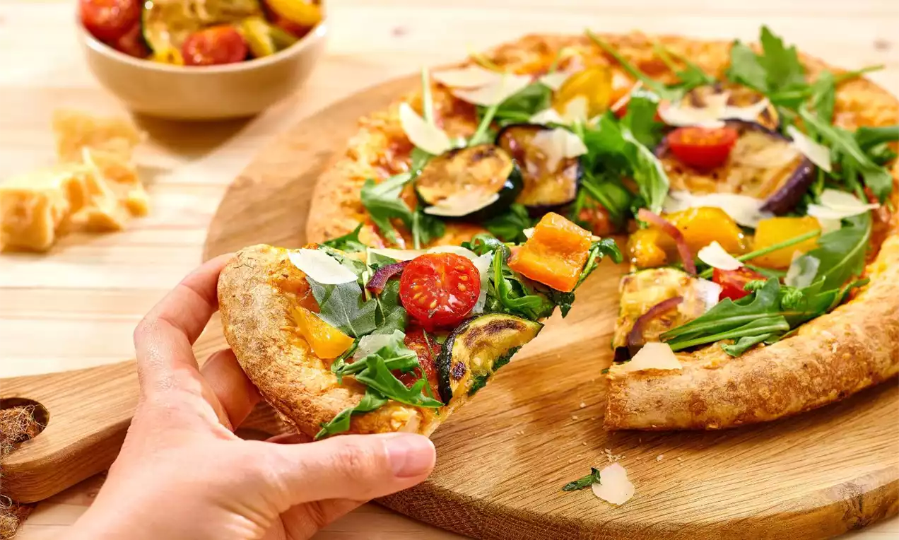 Pizza s křupavou zlatavou krustou, barevné kousky zeleniny na čerstvé rukole, drobné hoblinky sýra na dřevěném prkénku