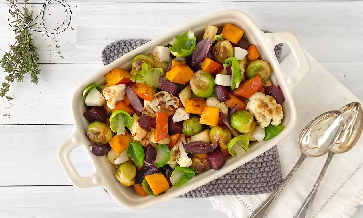 Buntes Ofengemüse mit goldbraunen Röstspuren aus Rosenkohl, Süßkartoffeln, roten Zwiebeln und Blumenkohl in heller Auflaufform auf Stoffunterlage