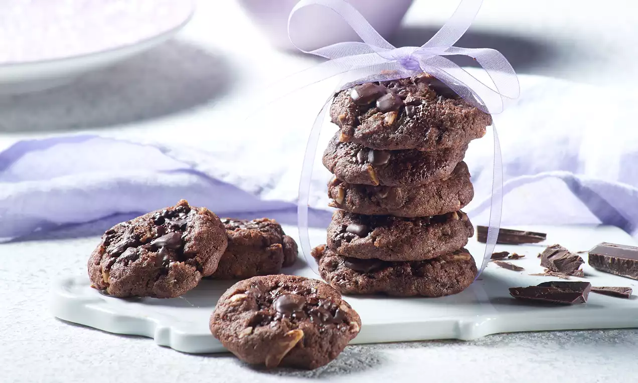 Tamnosmeđi veganski keksi okruglog oblika s nepravilnim površinama, komadićima čokolade i badema na bijelom pladnju uz ljubičastu vrpcu