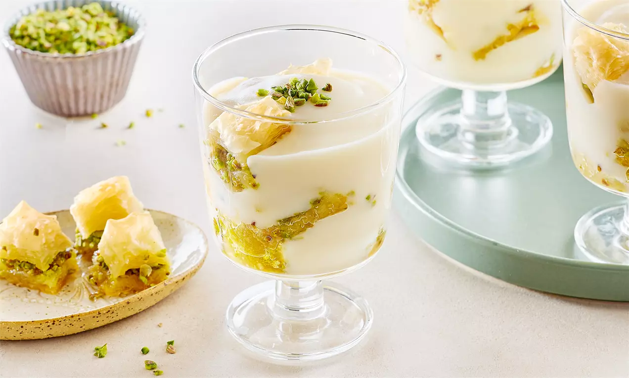Şeffaf kup bardağında katmanlı sunulan baklava parçaları, kremsi beyaz muhallebi ve ince kıyılmış yeşil fıstıkla süslenmiş