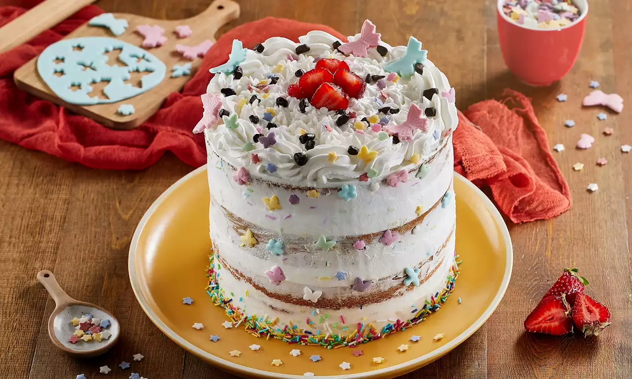 Kat kat beyaz kremayla kaplanmış naked cake, üstünde çilek ve renkli şekerlemelerle süslü, sarı tabakta ahşap masada sunulmuş