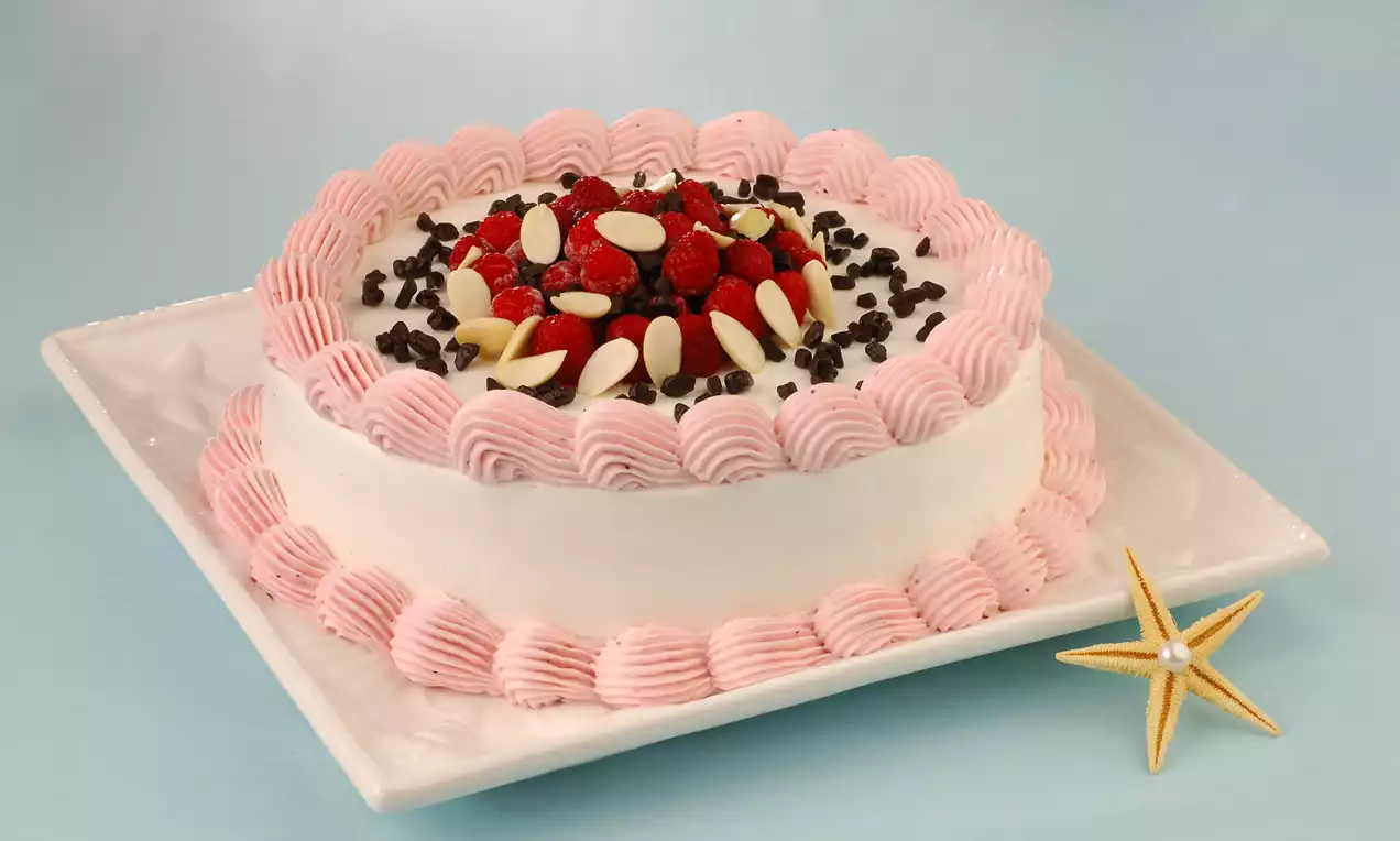 Yuvarlak beyaz tabakta, kenarları pembe krema ile süslenmiş, ortasında orman meyveleri, beyaz badem dilimleri ve çikolata parçaları bulunan pasta