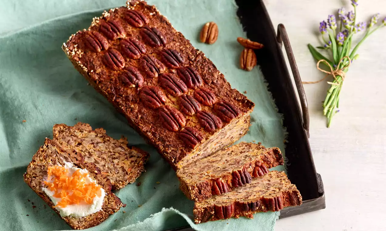 Rechteckiges, goldbraunes Eiweißbrot mit Pecannüssen belegt, eine Scheibe mit Frischkäse und Karottenraspeln auf grünem Tuch drapiert