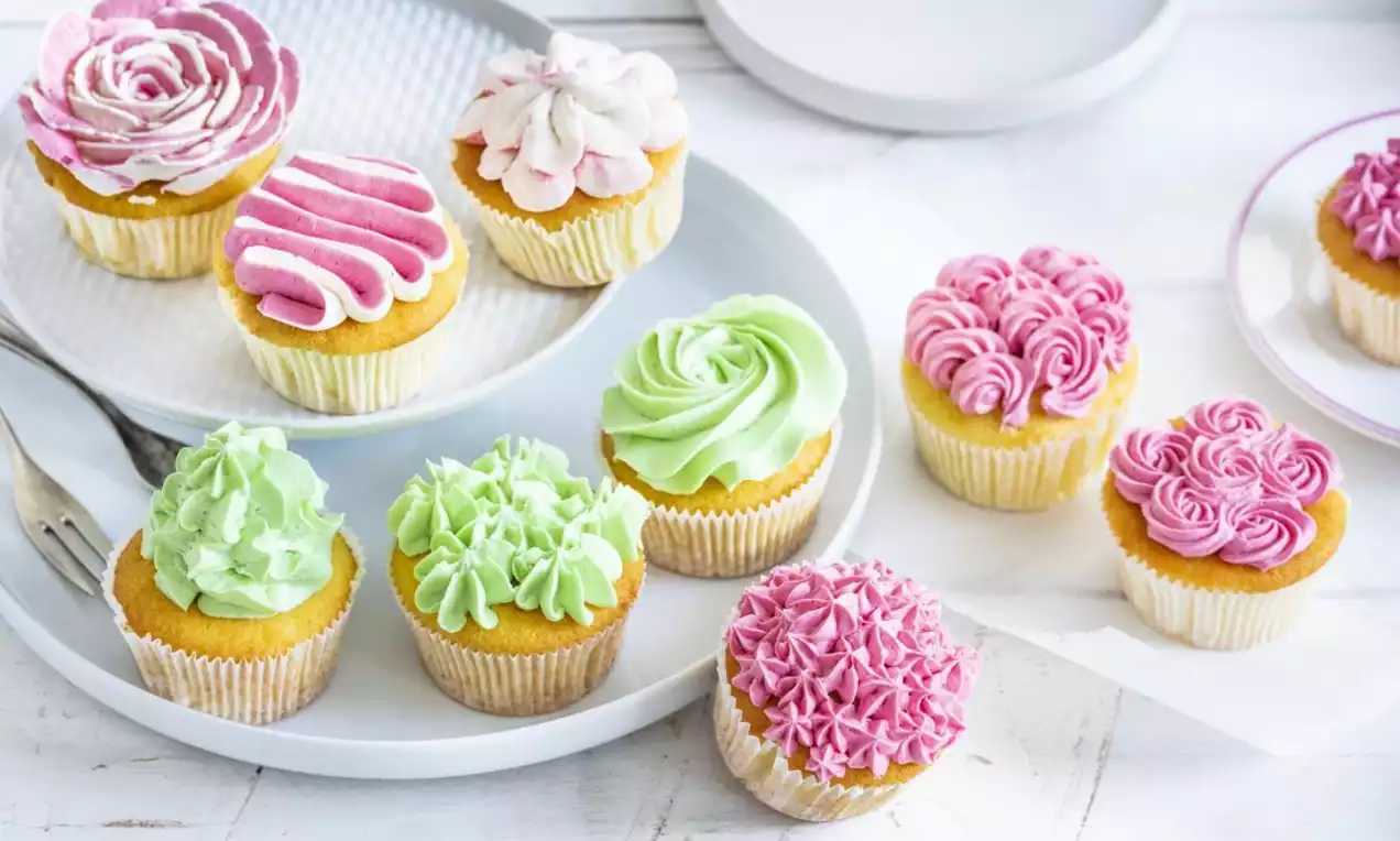 Golden cupcakes topped with pastel pink, green and white swirled frosting in floral shapes, arranged on white plates on a light wooden table