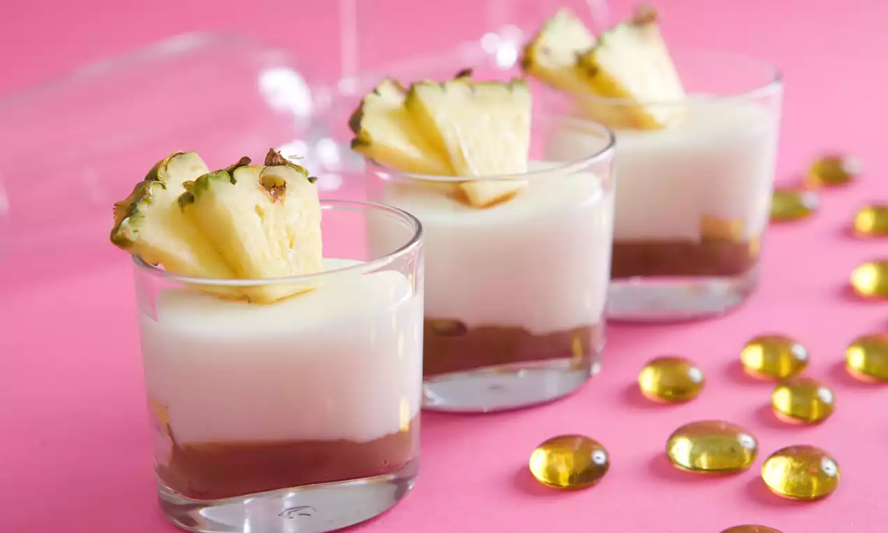 Skleničky s bílým kokosovým pudinkem na hnědé vrstvě, ozdobené plátky ananasu, na růžovém pozadí s dekorativními zlatými kamínky