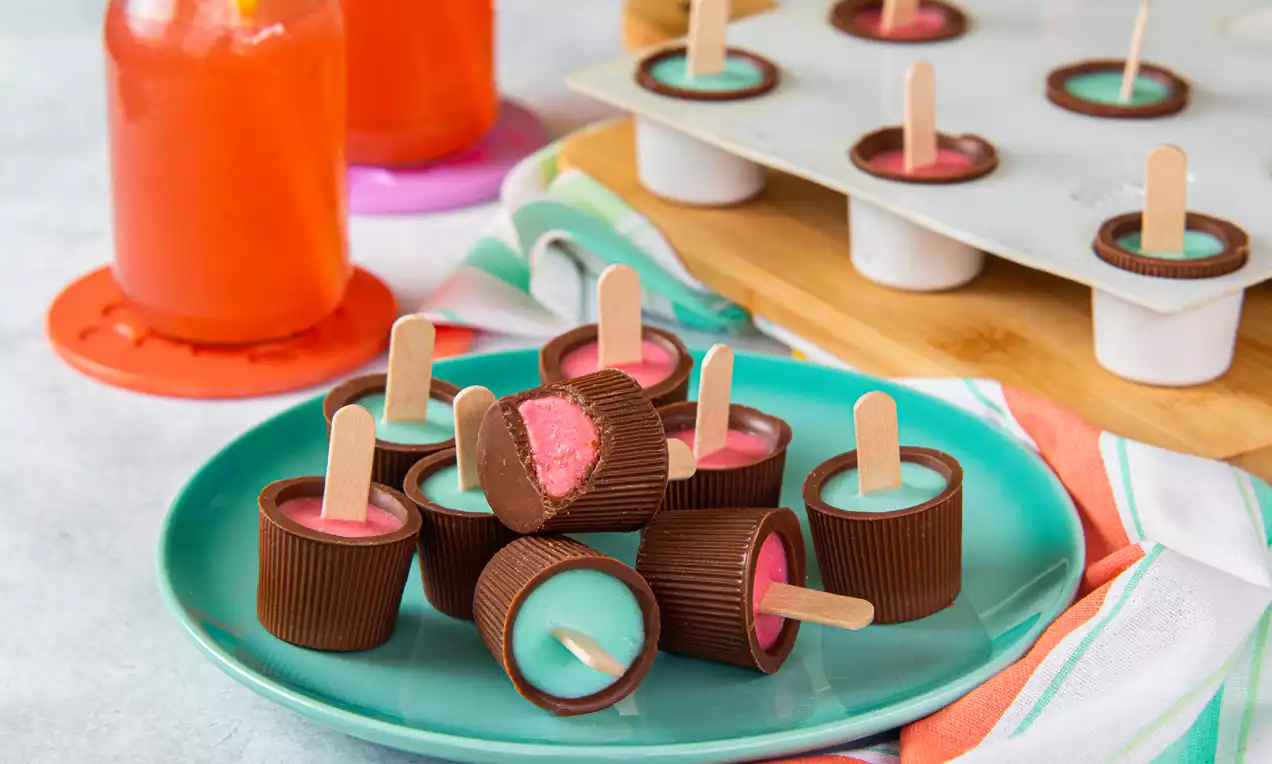 Copinhos de chocolate marrom recheados de gelatina rosa e azul, com palitos de madeira, dispostos em um prato azul com toalha colorida