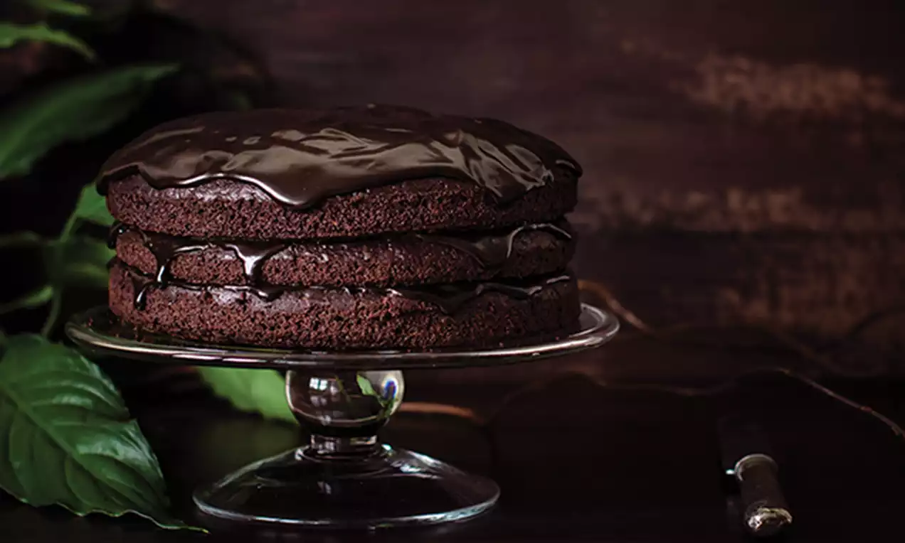 Rich, dark chocolate Devil's food cake with three moist layers, glossy ganache glaze, set on a glass cake stand against a wooden backdrop