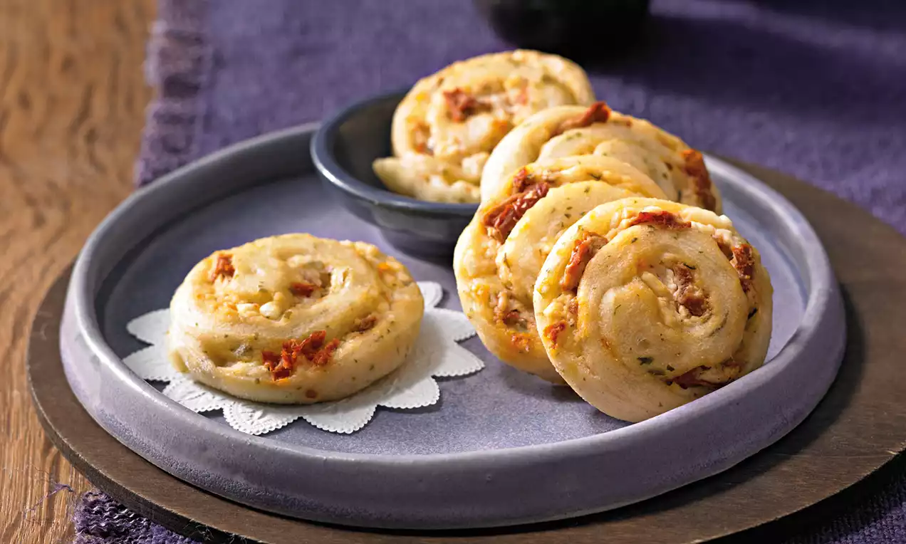 Goldgelbe, weich gebackene Hefeschnecken mit Kräutern und Tomaten auf weißem Unterleger, angerichtet auf einem lilafarbenen Teller