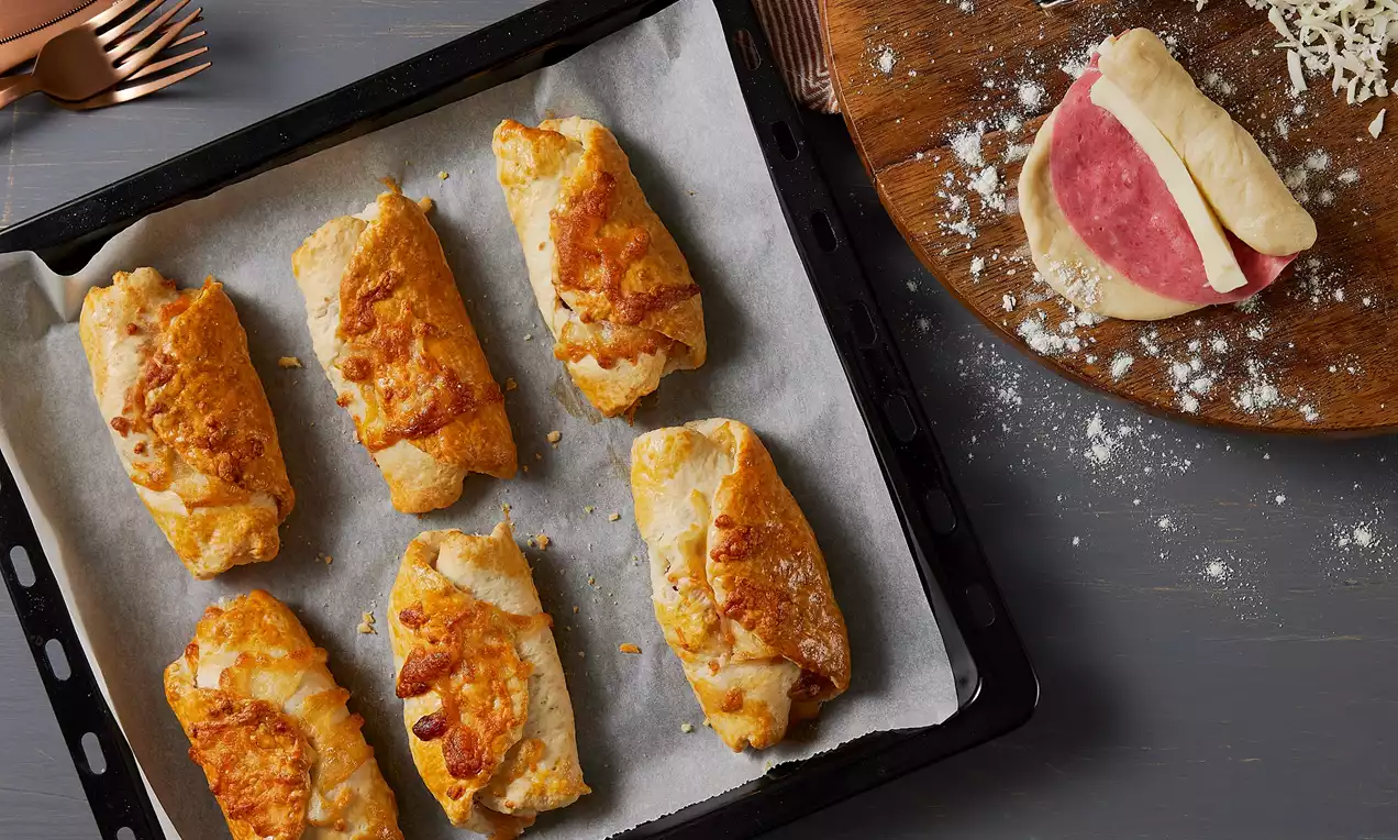 Altın rengi kızarmış salamlı ve peynirli çörekler, yağlı kağıt serili fırın tepsisinde, yanında tahta üzerinde hazırlık aşaması görülüyor