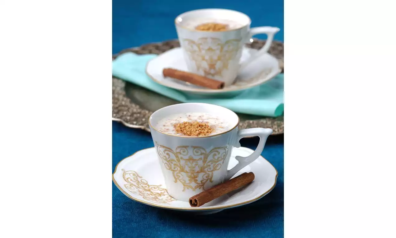 Altın desenli beyaz fincanda sunulan kremalı salep, üzerine serpilmiş tarçınla mavi zemin üzerinde, yanında tam bir tarçın çubuğu ile