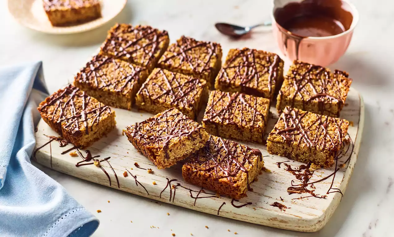 Golden flapjack squares with a crumbly texture drizzled with dark chocolate, arranged on a wooden board beside a copper bowl of melted chocolate
