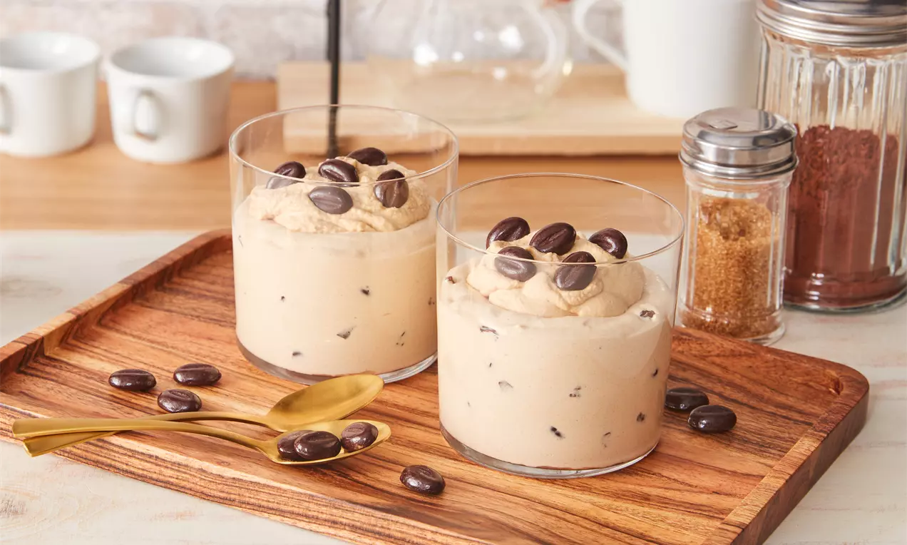 Zwei Gläser mit cremiger, beigefarbener Kaffeecreme, dekoriert mit dunklen Schokoladenkaffeebohnen auf einem Holztablett mit goldenen Löffeln