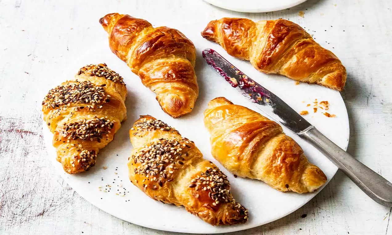 Croissant dorati e lucidi, alcuni cosparsi di semi, disposti su un piatto bianco con un coltello sporco di marmellata accanto