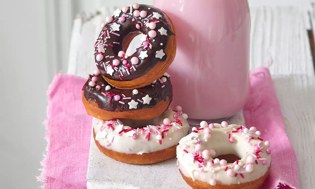 Kulaté donuty s čokoládovou a bílou polevou, zdobené růžovými, bílými kuličkami a hvězdami, na růžovém ubrousku vedle růžové láhve